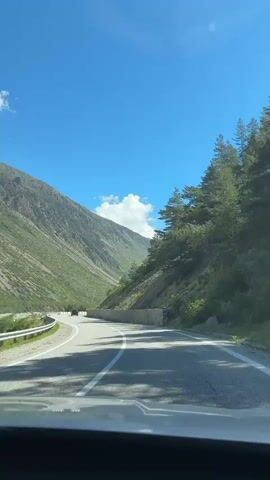 This is a first-person perspective video of driving on a paved road through a mountainous valley. The road curves to the left, with a steep, green mountain on the left and a rocky slope with trees on the right. The sky is a clear blue with a few white clouds. There are white lines marking the lanes of the road, and a low stone wall runs along the right side of the road. A few red flags on poles are visible along the left side of the road. The sun is shining, casting shadows on the road. The overall impression is of a scenic drive in a natural landscape.