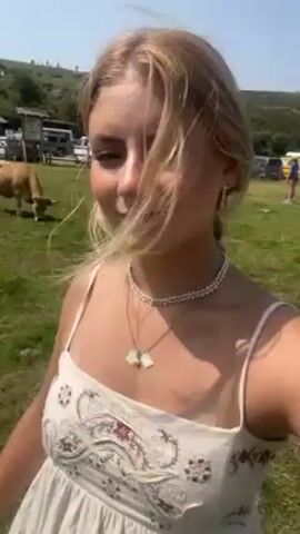 The video shows a young woman with blonde hair, wearing a white dress, a beaded choker, and a necklace, holding the camera and filming herself. She is outdoors in a grassy field, moving slowly and looking into the camera. In the background, there are several cows grazing, and in the distance, a parking area with several cars and vans is visible under a blue sky. The scene appears to be rural or agricultural.