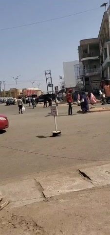The video shows a street scene in what appears to be an urban area in Africa. Cars and trucks are driving on the road. Pedestrians are crossing the street. Buildings line the street, and some appear to have businesses on the ground floor. The weather is sunny and clear. There are signs posted at crosswalks. There are overhead power lines.