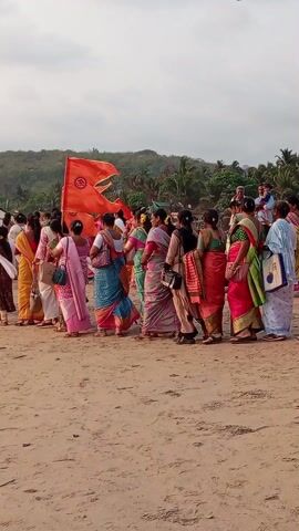 The video shows a large group of people, primarily women dressed in saris, walking together on a sandy beach. Many of them are carrying orange flags bearing what appears to be an emblem or symbol. The crowd appears to be moving away from the camera's perspective. In the background, there's a hill covered with greenery and trees, with some structures visible near the shoreline, suggesting a coastal town or settlement. The sky overhead is overcast. The general impression is that of a procession or rally taking place on the beach.