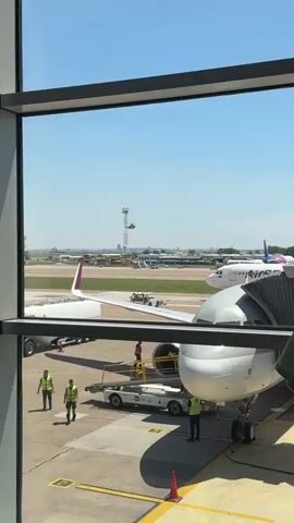 The video opens with a view of an airport tarmac through a window. In the foreground, the wing of a passenger jet is visible on the right, and part of the engine and fuselage are visible on the left. Several airport workers, wearing high-visibility vests and casual clothing, are seen around the aircraft. There are various pieces of airport equipment on the tarmac, including a baggage tug and a jet bridge. In the middle ground, another passenger jet's tail is visible. Beyond the tarmac, a tall radio tower stands with a helicopter in flight near its top. Further in the background, buildings and distant city skylines can be seen under a clear blue sky. The video then zooms in on the helicopter, which is a military-style helicopter, hovering in the air. The scene continues to focus on the helicopter as it moves across the sky, with the airport infrastructure and buildings remaining in the background.