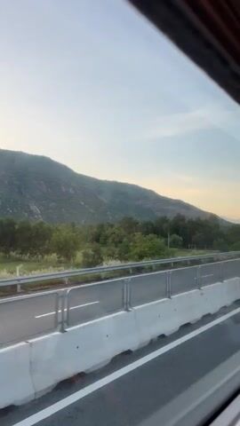 This media file captures a scenic drive along a highway in Công Hải, Vietnam, at what appears to be either dawn or dusk, judging by the soft, warm light illuminating the landscape. The primary subjects are the highway itself, with its clear white lane markings and concrete barriers, and the surrounding natural environment. Rolling green hills, covered in lush trees and vegetation, form a majestic backdrop to the road.

Several vehicles are visible on the highway, including a dark SUV and a red sedan, indicating normal traffic flow. The highway is lined with metal guardrails and concrete dividers, suggesting a well-maintained road infrastructure. Tall utility poles and a large road sign are also present, characteristic of a highway setting. The sky is a pale blue with hints of yellow and orange near the horizon, further reinforcing the impression of either sunrise or sunset. The overall mood is peaceful and serene, showcasing the beauty of the Vietnamese countryside from the perspective of a moving vehicle. There is no discernible text visible in the media file.
