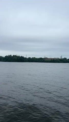 The video shows a wide body of dark, choppy water under a cloudy, overcast sky. In the distance, a tree-covered shoreline is visible. On the shoreline, there are several structures. Towards the left side of the frame, a tall, slender tower, possibly a lighthouse, stands out against the sky. Further to the right, there are other industrial-looking structures with cranes. The overall impression is a somewhat somber, industrial waterfront scene. The camera appears to be on a boat moving slowly through the water, as evidenced by the slight movement of the frame and the rippling water. There are no people visible in the video.