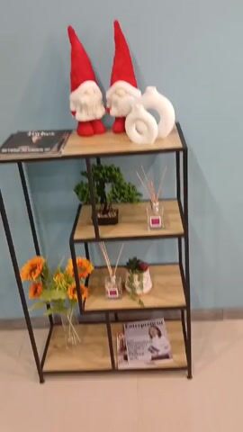 The video shows a four-tiered shelving unit with a dark metal frame and light wood-look shelves. The top shelf contains a copy of Forbes magazine, two gnome-like Christmas decorations with red hats and white beards, and two white ceramic vases. The second shelf from the top holds a small decorative bonsai tree and a glass bottle with diffuser reeds. The third shelf contains another glass bottle with diffuser reeds and a small potted succulent. The bottom shelf contains a copy of Entrepreneur magazine and a glass vase holding orange sunflowers. The shelving unit is placed against a light blue wall and on a light brown tiled floor. The camera provides a static shot of the shelving unit.