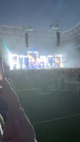 The video opens with a view of a large outdoor concert stage at dusk or night, illuminated by numerous bright lights and spotlights. Towering speaker stacks are visible on either side of the stage, supported by metal scaffolding. The stage itself features a massive LED screen displaying abstract blue patterns and geometric shapes, with bright white lights forming large block letters that appear to spell out "OTP." In front of the screen, there are smaller light effects and possibly pyrotechnics, with flames erupting at one point. A large crowd of people is gathered in front of the stage, visible in silhouette due to the bright stage lighting. Some individuals in the crowd are holding up their phones to record the performance. The overall atmosphere is energetic and celebratory, typical of a large music event. The video appears to be shot from within the audience, looking towards the stage.