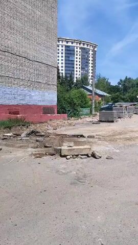 The video shows a construction site. In the foreground, there is a paved area with rubble and broken concrete. In the middle ground, a yellow backhoe loader is parked on a dirt area. There are stacks of concrete blocks next to the backhoe. In the background, there are several apartment buildings, trees, and a blue sky with white clouds. The overall scene depicts an urban environment undergoing construction work.