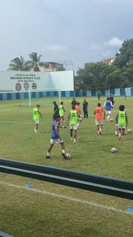 A daytime outdoor wide shot shows a group of about twelve people, mostly men, in an open field, wearing athletic attire and playing with soccer balls. Some men are in neon green sleeveless shirts, some in orange sleeveless shirts, and some in blue sleeveless shirts. A few people wear white shorts, while others wear black shorts. One man is wearing black pants and a black short-sleeved shirt. Some men are practicing dribbling the ball, while others are standing around. Some men are walking. Blue and white stripes are visible in the background, along with a building and many trees. The sky is partly cloudy. The grass is green.