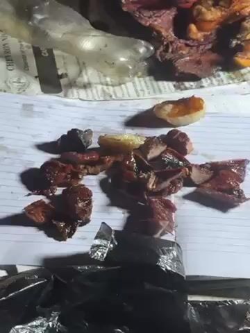 A person slices cooked pieces of meat and onions with a knife. The meat and onions are placed on white lined paper. There is more meat to the top of the paper, with some of it also sliced. There is some plastic and foil nearby. The person has dark skin. The lighting appears dark.