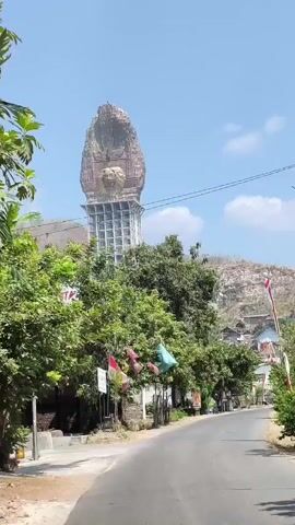 The video shows a wide shot of a street with a large, imposing statue of a face at the end. The statue is weathered and appears to be made of stone, with intricate carvings and a distinctive headdress. It sits atop a tall, modern-looking structure with scaffolding. The street is paved and curves slightly to the right, lined with trees and buildings. Red and white flags are visible, suggesting a national holiday or celebration. The sky is clear and blue. The overall impression is one of a significant landmark in a rural or semi-urban setting.
