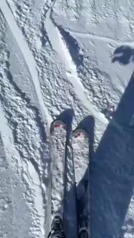 The video shows a person skiing down a snowy slope on a sunny day. The camera appears to be mounted on the skier, showing a first-person perspective of the descent. Initially, only the skis and the snowy terrain are visible. The view then widens to show other skiers and snowboarders on the slope, along with snow-covered mountains in the background.  The skier briefly turns the camera towards themselves, revealing a person wearing a helmet and goggles, smiling and speaking. The bright blue sky is visible above the mountain range.