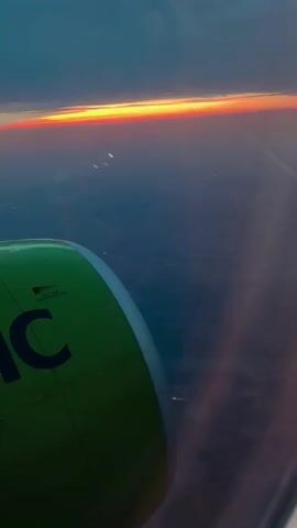 The video shows a view from an airplane window during what appears to be dusk or dawn. The foreground is dominated by a portion of the aircraft's green engine nacelle, with the partial logo "IC" visible. Beyond the wing, the horizon displays a vivid band of orange and yellow light from the setting or rising sun, set against a gradient of blue and purple sky. Below the horizon, the landscape is dark, with scattered pinpricks of light suggesting civilization. The overall impression is one of aerial travel at a transitional time of day.