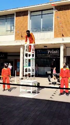 A performer in a bright orange and yellow jumpsuit stands precariously atop a tall, unstable tower constructed from white, open-sided modules. Below the performer, the tower is comprised of three stacked sections, each with a slightly different configuration of vertical and horizontal supports, creating a series of open cubbies. The tower rests on a white rectangular platform, which in turn is supported by two white, L-shaped legs. To the left of the tower, another performer, also in an orange and yellow jumpsuit, stands facing the tower. To the right of the tower, a third performer, similarly dressed, stands facing away from the tower, looking towards the right. A black cord runs horizontally across the foreground, behind the performers. In the background, a brick building with large windows rises above the scene. A sign for "LiverPrinti School Uniform, Workwear & Printing" is visible on the building. Several spectators are scattered around the area, observing the performance. The ground is paved with dark bricks. The sky is clear and blue, indicating a sunny day.