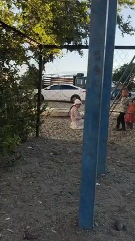 The video shows a scene outside, likely in Balykchy, Kyrgyzstan. In the foreground, a chain-link fence is visible, with a dirt and gravel ground. Beyond the fence, several children are present. One child, wearing a white hat and a light-colored outfit, is sitting down. Another child, wearing a pink dress and a pink hat, is also seated. Further back, two other children are standing, one in a light-colored shirt and dark pants, and the other in a red dress. A white sedan and a dark-colored truck are parked in the background. There is also a swing set visible, with a person, possibly a child, on top of it. Trees with green foliage are present, and the sky is clear blue, suggesting it is daytime and the weather is pleasant. The overall impression is that of children playing outdoors in a somewhat rustic setting. There are no visible text elements in the video.