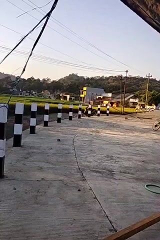 The video shows an outdoor scene at what appears to be a rural or suburban intersection. The foreground is a concrete road with a thin, dark line running down its center, possibly a joint or crack. On the left side of the road, there is a row of black and white striped bollards. In the background, there is a rice field to the left, and several buildings and houses to the right, situated on a gentle slope. Trees and vegetation are visible on the hills in the far background. Power lines crisscross the sky. The lighting suggests it is either early morning or late afternoon, with a warm, soft light. A bicycle is parked on the right side of the road, and a person is visible in the distance walking or standing near the buildings. The overall impression is a quiet, transitional space between cultivated land and a residential area.