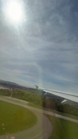 From the perspective of an airplane window, the viewer sees the plane’s wing extended over a green, grassy landscape dotted with trees and a cityscape. The bright sun shines through a sky filled with scattered clouds. A mountain range is visible in the distance. The plane appears to be in flight.