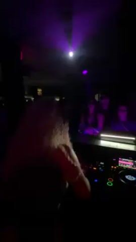 The video shows a DJ performing at a club. The DJ, a woman with curly blonde hair, stands behind a DJ console illuminated by a small lamp. The console includes a Pioneer DJM-900NXS2 mixer. A purple spotlight shines on the DJ and the crowd. A group of people, some wearing sunglasses, is visible in front of the DJ. The club is dark with minimal lighting. The DJ is actively mixing music. The crowd seems to be enjoying the music. The atmosphere is energetic and party-like.