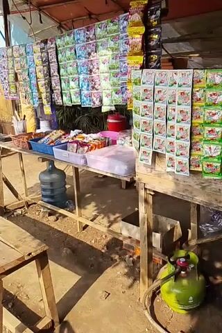 The video shows an outdoor market stall with a variety of goods displayed on wooden tables. The stall is covered by a reddish awning. A large blue water jug is visible beneath one of the tables. The tables are laden with packaged snacks, drinks, and other small items. A gas cylinder is positioned on the ground near the right side of the stall. A motorcycle is partially visible on the left side of the scene. The ground appears to be dirt or gravel, and sunlight casts shadows across the area, suggesting it is daytime.