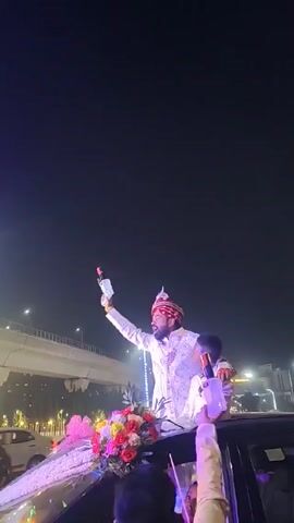 At night, a groom in a white and pink sherwani and a turban stands on the roof of a black car. He holds up a noise maker in his left hand and a small boy stands to his side, holding a similar noise maker. The car is decorated with pink and red flowers on the hood. A large, illuminated, frilled umbrella is being raised above the pair. The car is in a procession with other people nearby. Street lights and an overpass are visible in the background.
