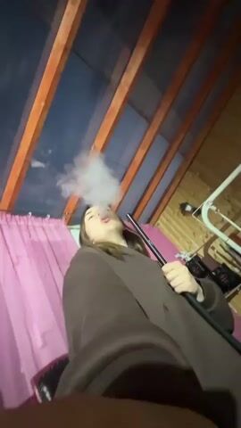 A woman with long brown hair is holding a hookah hose and exhaling smoke in a room with wooden walls and ceiling. She is wearing a brown robe-like garment. There are pink curtains to her left and various objects are visible in the background, including what appears to be medical equipment. The camera angle is low and tilted upwards.