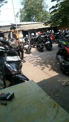 The video shows an outdoor parking lot filled with many motorcycles. Two people are visible in the center of the frame, with one walking and the other standing. The ground is paved and appears to be dusty. Trees with green foliage are present in the background, providing shade. The lighting suggests it is daytime, possibly sunny. Some of the motorcycles are red, black, and green. One of the people is wearing a backpack.