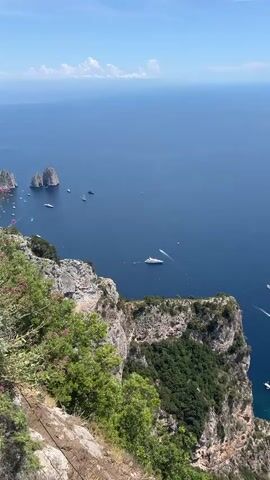 An aerial shot shows a coastline with steep cliffs covered in greenery. The azure sea below is dotted with numerous boats, including a large yacht and several smaller vessels. The sky is clear and blue with a few wispy clouds on the horizon. The camera pans slightly, revealing more of the coastline and the vast expanse of the ocean.