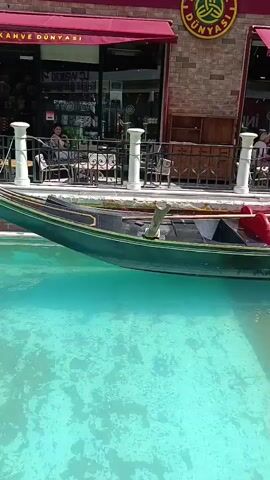 A gondola floats in a turquoise canal. Behind the gondola, there is a building with a red awning and brick facade. A sign on the building reads "KAHVE DÜNYASI" in English and Turkish. People are seated at tables outside the building, some are drinking and eating. The water in the canal is clear and blue, with the bottom visible beneath the surface. The gondola is dark green with red seats and a wooden oar. The sun is shining, casting shadows on the water and building.