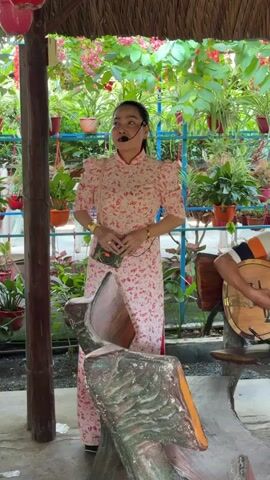 The video captures an outdoor musical performance under a thatched roof, likely in Thới Sơn, Vietnam. A woman in a pink floral ao dai and a microphone headset stands and sings, holding a small green bag. She wears gold bracelets on both wrists. Beside her, a man sits and plays an acoustic guitar, accompanying her. Another man is partially visible at a wooden table with a white bowl on it. The setting is vibrant with numerous potted plants, featuring both green foliage and red flowers, creating a lush garden atmosphere. The scene is brightly lit, suggesting daytime.