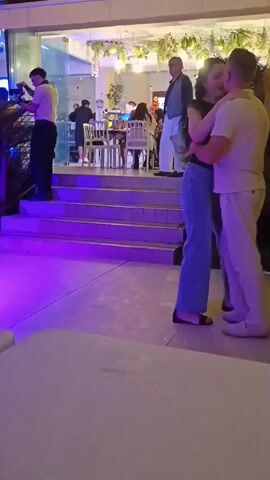 A man and woman dance together at an outdoor event. The man wears a light-colored shirt and pants. The woman wears a dark top and blue jeans. They are in the foreground, facing away from the camera. Behind them, a set of stairs is illuminated with colored lights, shifting from green and red to blue and purple. Several other people are visible in the background, some seated at tables and others standing. The environment appears to be a restaurant or event venue with outdoor seating. The lighting suggests it is nighttime or evening.