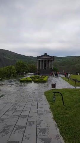 The video shows the Temple of Garni in Armenia on an overcast day. The temple is a classical Greco-Roman style building with columns. People are walking around the temple and in the surrounding area. The grounds are wet, suggesting it may have recently rained. A stone pathway leads towards the temple, and there is grass on either side of the path. There are hills in the background. A small garbage can and a lamppost are visible on the right side of the pathway.