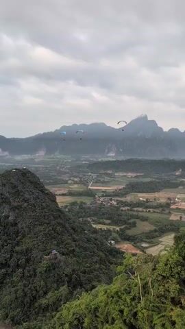 The video shows a panoramic view of a lush green valley with mountains in the background. Several paragliders are visible in the sky, soaring over the landscape. The foreground is dominated by a steep, forested hillside, with some buildings or structures visible nestled amongst the trees. The valley floor is a patchwork of fields, possibly rice paddies, with scattered villages and roads. The sky is overcast with dramatic clouds, casting a soft light over the scene. The overall impression is one of natural beauty and adventure.