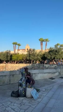 A person is playing the cello on a paved walkway. A cello case and a backpack are next to them. A few children are watching. Palm trees and a building are in the background. The sky is clear and blue.