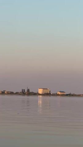 A wide shot of a body of water under a light blue and pink sky. A shoreline is visible in the distance, dotted with several buildings of varying sizes and architectural styles. The largest building in the center of the frame is a light-colored, multi-story structure with a flat roof and numerous windows, casting a reflection on the calm water. To its left, a tall, slender structure with a pointed top, resembling a minaret or a church steeple, stands out against the sky. Other smaller buildings line the shore to the left and right of these prominent structures. The water surface is rippled, catching the soft light of the sky. No people or animals are clearly visible. The overall impression is a serene, coastal or lakeside town scene during twilight or dawn.