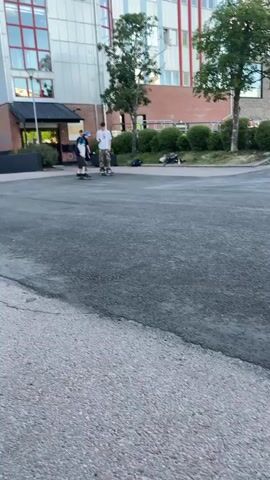 The video shows a group of young males skateboarding in an asphalt area outside a brick building. Two of the males are closer to the camera, with one performing a trick on a skateboard. Another male is standing behind him, observing. Further in the background, other males are visible on skateboards, and some are standing. There are trees and bushes in the background, as well as a car parked near the building. The lighting suggests it is daytime.