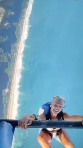 A person in a skydiving suit jumps from a platform over a body of water and a coastline. The person is smiling and holding onto the edge of the platform before letting go. The camera is attached to the person, providing a first-person perspective.