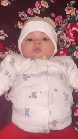 A baby lies on its back on a floral patterned surface. The baby wears a white quilted jacket with teddy bear and cloud designs, red pants, and a white and beige hat. The background is a dark fabric with large pink and red flowers and green leaves. The baby is looking directly at the camera.