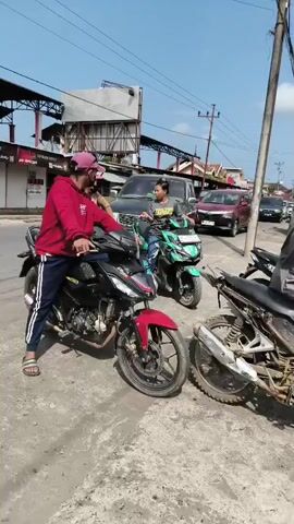 The video shows a street scene with multiple motorcycles and cars. In the foreground, two motorcycles are visible, one red and black, and another mostly black and rusty. A person in a red hoodie and a pink hat is sitting on the red and black motorcycle. Another person in a grey shirt and a bucket hat is on the rusty motorcycle. In the background, a green scooter is parked, and a person is sitting on it. Several cars and trucks are also present, and a gas station sign is visible. The overall environment appears to be a busy street on a sunny day.