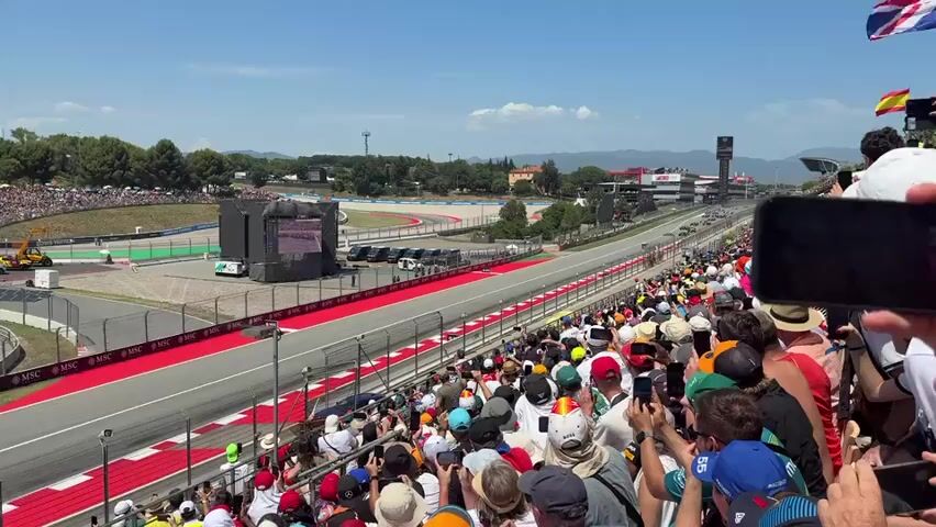 The video captures a Formula 1 race from a spectator's perspective in a grandstand. The camera is positioned to show a wide view of a curving race track section, with many spectators filling the seats in the foreground. Multiple Formula 1 cars are visible on the track, some in the distance and others closer to the camera, with their distinctive aerodynamic shapes and sponsor liveries. The track itself has a mix of grey asphalt and red and white curbing. Beyond the track, there are grassy hillsides densely populated with more spectators, and further in the background, trees and buildings are visible under a clear blue sky. A large screen is positioned trackside, displaying images or data related to the race. Various vehicles, including what appears to be a safety car or support vehicle and an excavator, are present in the pit or off-track areas. The spectators are diverse in age and gender, many holding up smartphones to record or photograph the action. Some are wearing hats, and their clothing varies in color and style. The overall atmosphere suggests an exciting and well-attended sporting event.