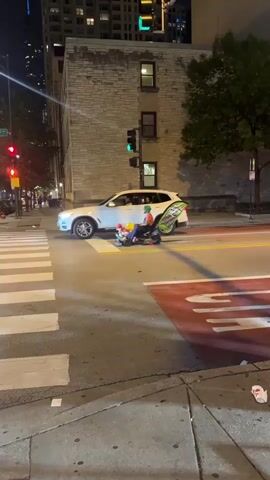 At night, in an urban intersection, a person sitting in a motorized chair faces away from the camera while stopped within a crosswalk. The person is wearing an orange helmet and a green reflective vest. The chair is decorated with rainbow-colored pinwheels and a green sign is attached to the back of the chair. A white SUV is stopped directly in front of them, facing the same direction. Both the SUV and the motorized chair are stopped at a red traffic light. Pedestrians stand on the sidewalk in the background. Tall buildings are visible in the distance. A street sign reading "Chicago St" is on a pole to the left. The scene is lit by streetlights and traffic signals.