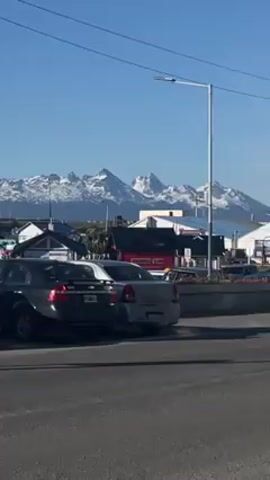 The camera pans horizontally across a street scene with a backdrop of snow-capped mountains under a clear blue sky. In the foreground, there's a row of parked cars along the roadside. Beyond that, there are rooftops of houses and smaller buildings, with a busy street intersection. The camera continues to pan towards the right, revealing larger boats and yachts docked at a harbor. A pedestrian is visible near the waterfront, and vehicular traffic moves along the street.