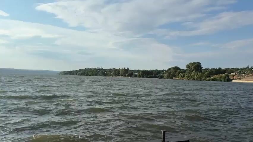 The video shows a wide expanse of choppy water under a bright, partly cloudy sky. The sun is visible on the left side of the frame, its rays reflecting off the water's surface. The shoreline is visible in the distance, lined with trees and some buildings. The camera appears to be mounted low, possibly on a boat or a dock, as the waves are close to the bottom of the frame. There are no people or animals visible in the video. The overall impression is one of a natural landscape on a sunny day.