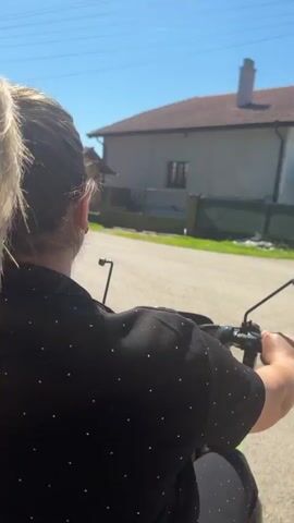 The video shows a first-person perspective of someone riding a motorcycle on a rural road. The rider's blonde hair is visible, pulled back into a ponytail. They are wearing a black shirt with white polka dots. The road is paved and appears to be in a residential area, with houses and trees lining the sides. The sky is clear and blue, indicating a sunny day. A red van is parked on the left side of the road. The rider's hands are on the handlebars, and they are wearing a ring on their left hand. The motorcycle appears to be moving at a moderate speed.