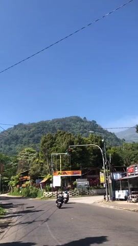 The video shows a rural road with a clear blue sky. There are trees and mountains in the background. On the right side of the road, there are several buildings with signs. Two people on motorcycles are driving on the road. The traffic light is red.
