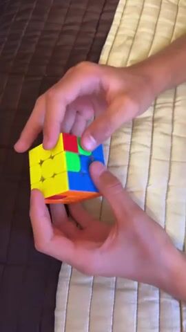 This is a close-up, top-down video of a person's hands manipulating a Rubik's cube. The cube is mostly yellow on one side, with some blue and green sections visible. The hands are turning and twisting the cube, but the exact moves are difficult to discern due to the speed and blur. The background appears to be a bed with a brown and beige striped blanket.