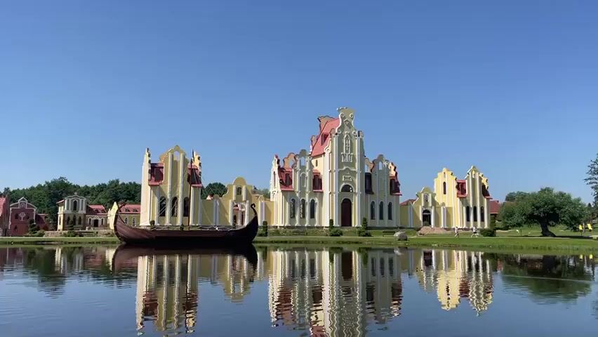 The video opens with a wide shot of a yellow and red building complex reflected in a lake. A long, narrow boat is docked on the left side of the frame. The sky is clear and blue. The building complex appears to be a series of connected structures with ornate architecture, featuring prominent red roofs and white trim. The reflection in the water is clear and undisturbed. The environment appears to be a park or estate with trees visible in the background. The camera is stationary.