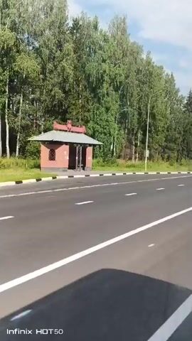 A dashcam video captures a view of a paved road from a moving vehicle. The road has white lane markings. On the left side of the road, there is a bus stop shelter and a dense line of trees, primarily consisting of deciduous trees with some coniferous trees interspersed. The sky is partly cloudy with blue sky visible. Several vehicles are seen traveling on the road. At 00:02, a white panel van drives past. At 00:03, a grey sedan drives by, and a white vehicle is seen in the distance to the left. The video is timestamped at the bottom left corner.