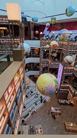 An overhead, high-angle, slow-pan shot captures a vast, multi-level bookstore. The architecture features tiered seating areas with people seated and walking. Bookshelves are visible on multiple floors, lining the walls and forming staggered structures. Suspended from the ceiling are numerous large, colorful spherical decorations resembling planets, some appearing to be planets with rings. A large yellow globe with colorful continents is prominently displayed in the center. Other decorative spheres, some resembling globes and others more abstract, are also present. The lighting appears to be natural light from a skylight above and artificial lights integrated into the shelving and seating areas, creating a warm and inviting atmosphere.