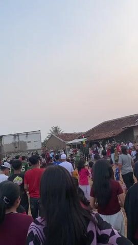 The video shows a gathering of people in an outdoor setting, possibly a village or rural area, with several buildings in the background. The sky is a pale orange, suggesting it might be sunset or sunrise. Many people, including children and adults, are standing and walking around, some in groups. There are hints of activities or events taking place, with some individuals appearing to be watching something or participating in a celebration. The ground is dirt and uneven. Flags are visible, including what appears to be an Indonesian flag. The atmosphere seems lively and communal.