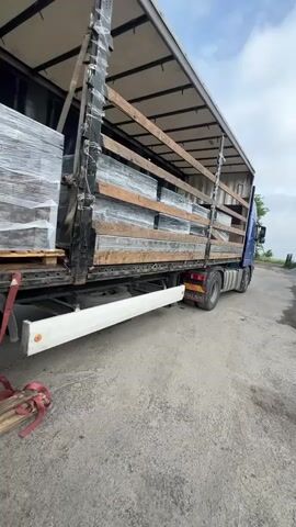 A blue and black Volvo semi-truck with a curtain-sided trailer is parked on a concrete or asphalt surface under a bright, partially cloudy daytime sky. The right side of the trailer is open, revealing its cargo.

Inside the trailer, several pallets of what appear to be building blocks or pavers are stacked and neatly arranged. Each pallet is shrink-wrapped in clear plastic, and some handwritten numbers, including "600," are visible on the wrapping of the leftmost pallet. The cargo is secured by multiple horizontal wooden slats that run along the length of the trailer, acting as load restraints.

On the ground directly beside the truck, a red tie-down strap is coiled, and a bundle of wooden poles or planks, also secured with a red strap, lies nearby. The truck features a white side underrun protection bar, which has partially visible text, possibly "CARGOTECH." The "VOLVO" logo is clearly visible on the truck's rear mudflap. In the distant background to the right, some greenery and trees can be seen.

The scene suggests that the truck is either in the process of being loaded, unloaded, or prepared for transport at a yard or delivery point in Uhry, Ukraine. No people are visible.