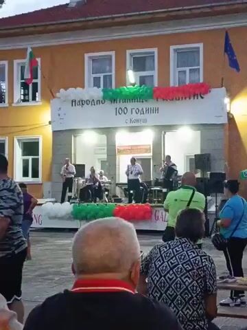 A live music performance is taking place outdoors in front of a building. A banner above the performance reads "народно читалище развитие 100 години с Кошуи". The building is yellow with orange trim, and has two floors with windows. Bulgarian flags and an EU flag are visible on the building. A stage is set up with musicians playing instruments, including a saxophone and an accordion. Several people are watching the performance from a courtyard area in front of the stage. The crowd includes men, women, and children. Some people are standing, while others are sitting at tables. Balloons in the colors of the Bulgarian flag are decorating the stage. The time is late afternoon or early evening.