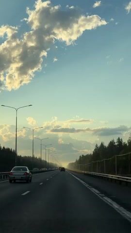 The video shows a view from a car driving down a highway. On the left side of the highway are streetlights and cars. On the right side of the highway are trees and a fence. The sky is blue with clouds. The sun appears to be setting or rising, casting a warm glow on the clouds and trees.